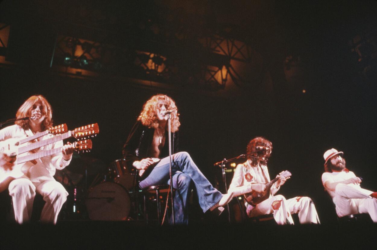 Led Zeppelin members John Paul Jones, Robert Plant, Jimmy Page, and John Bonham sit at the front of the stage during an acoustic portion of a 1977 concert.