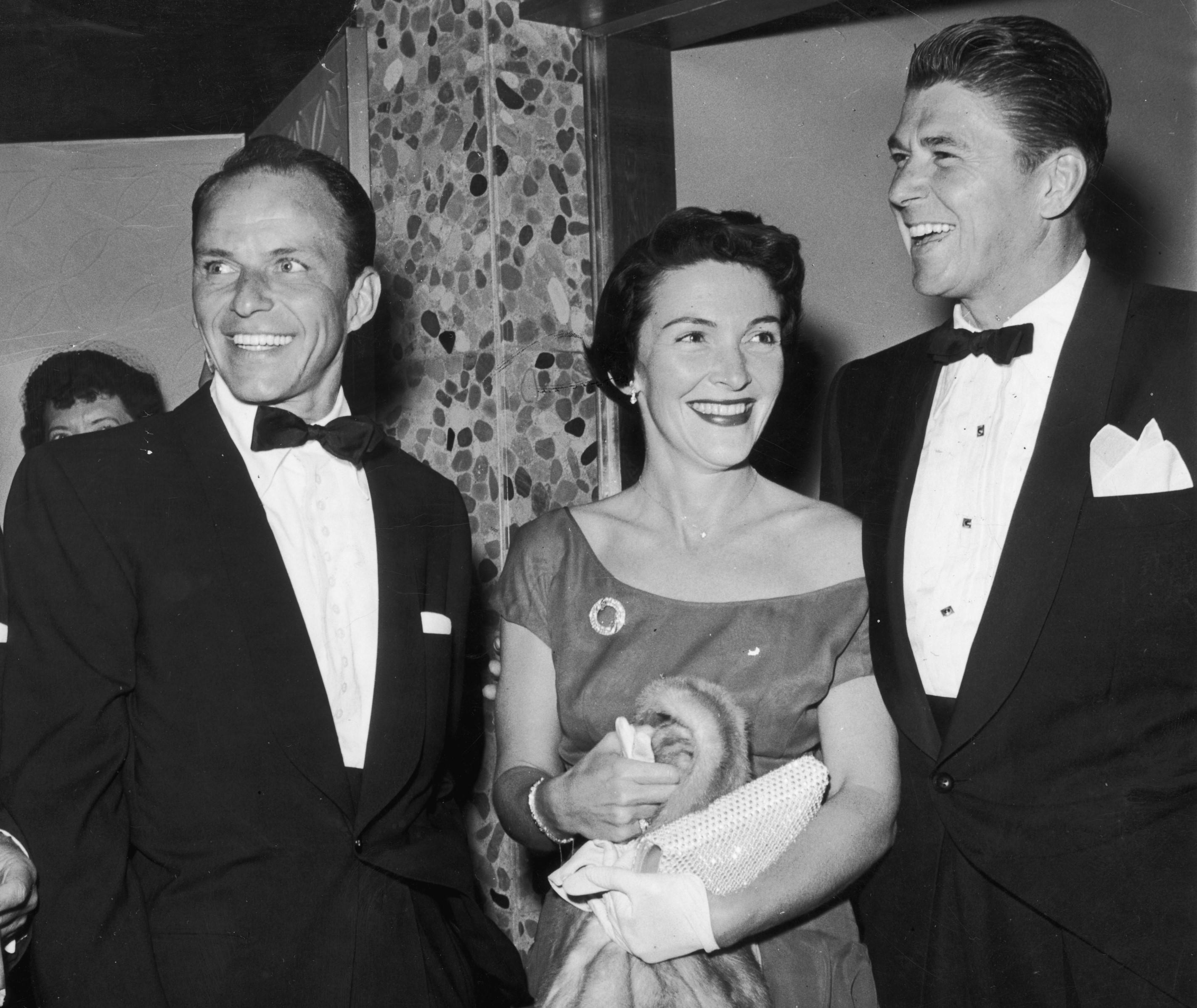 A black and white photo of Frank Sinatra, Nancy Reagan, and Ronald Reagan in 1955. They stand in a row and smile.